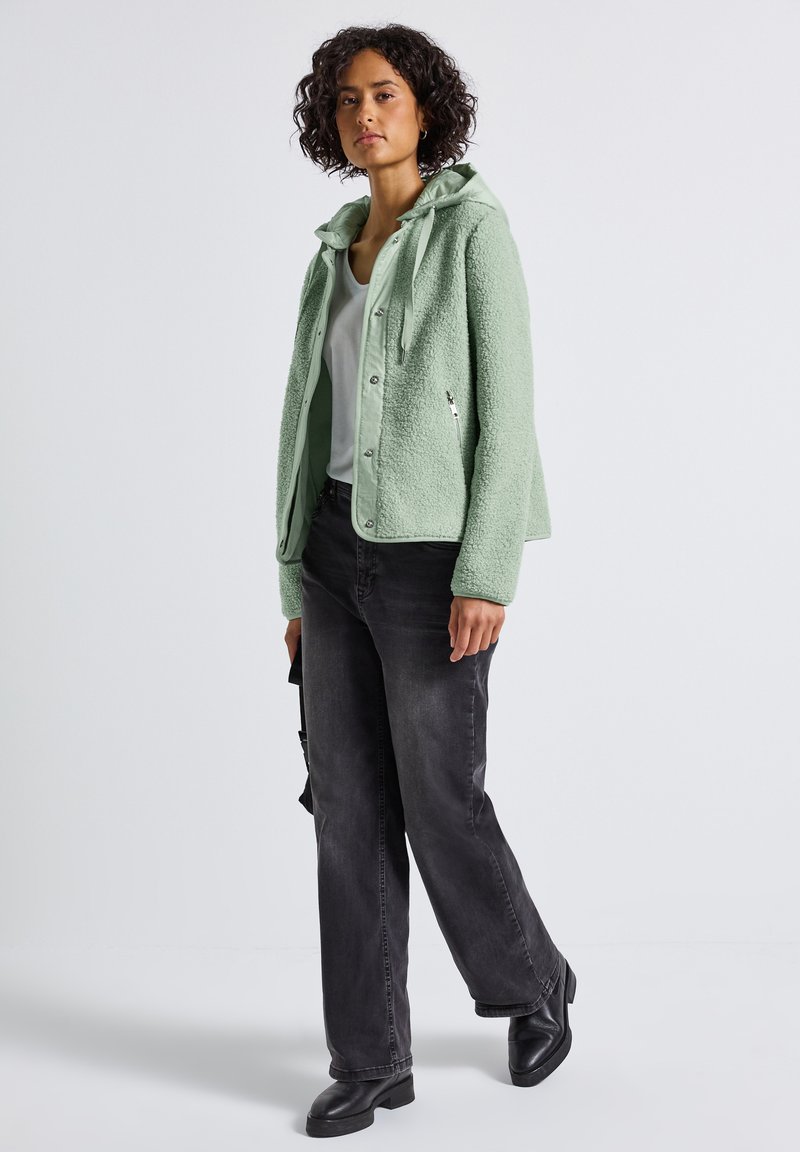 Donna in piedi con una giacca in pile verde chiaro, maglietta bianca, jeans a gamba larga scuri e stivali neri contro uno sfondo semplice.