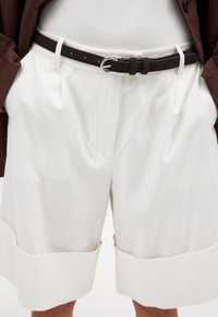 Hvide linnedshorts med en oplagt kant, der har frontfolder og et brunt ruskindsbælte med en sølvspænde. Glat tekstur og minimalistisk design.