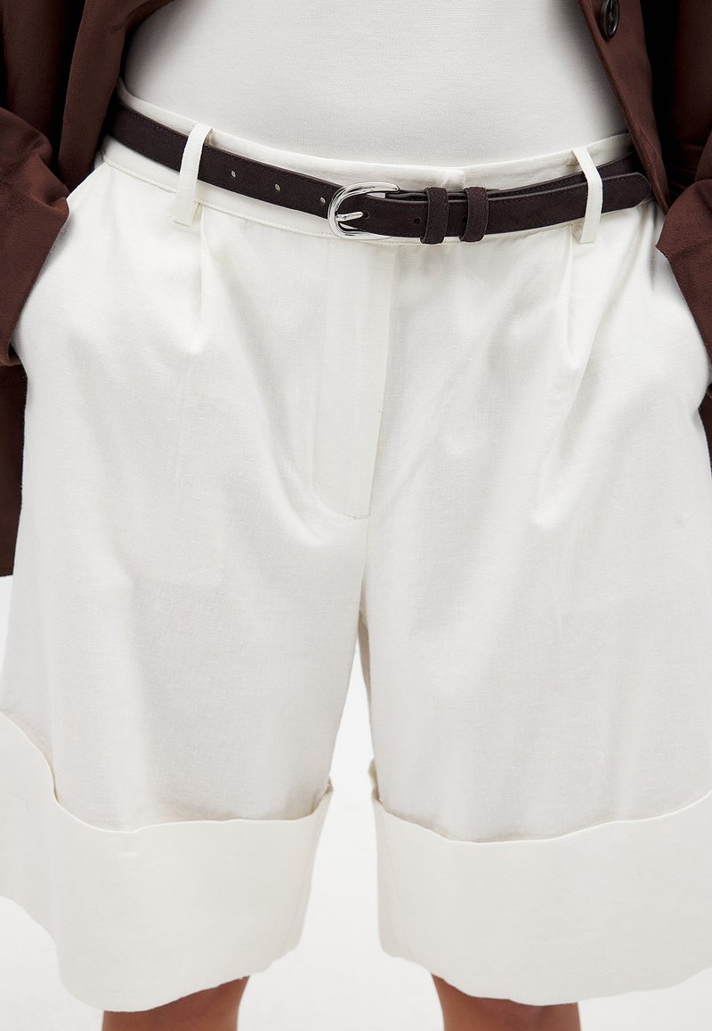 Hvide linnedshorts med en oplagt kant, der har frontfolder og et brunt ruskindsbælte med en sølvspænde. Glat tekstur og minimalistisk design.
