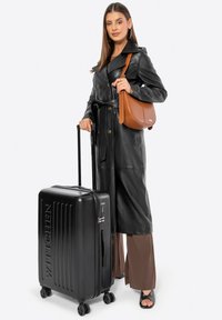 Maleta negra rígida con textura estriada, ruedas giratorias y asa superior. Modelo con abrigo de cuero negro y bolsa de crossbody marrón.