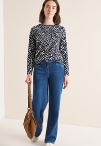 Navy lange mouw top met een wit abstract patroon, gecombineerd met lichtblauwe rechte jeans. Draagt een bruine suède tas met een korte strap.