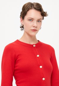 Cardigan rouge en tricot texturé, avec un col rond et cinq boutons dorés sur le devant. Manches longue avec une coupe ajustée.