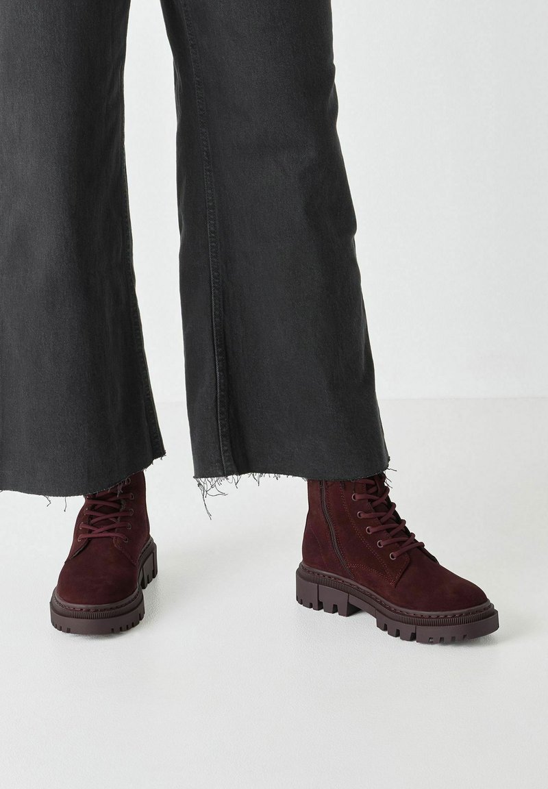 Bottines en daim bourgogne avec fermeture lacée à l'avant, semelle en caoutchouc épaisse et embout renforcé ; assorties à un pantalon noir à jambes larges avec un ourlet effiloché.