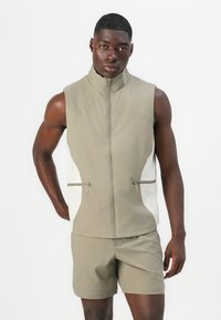 Sleeveless light brown athletic vest with high collar, front zipper, white side panels, and zippered pockets. Smooth texture, fitted design.