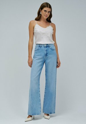Frau trägt weiße Spitzen-Camisole, hellblaue Jeans mit hoher Taille und weitem Bein sowie weiße spitze Schuhe, steht vor grauem Hintergrund.