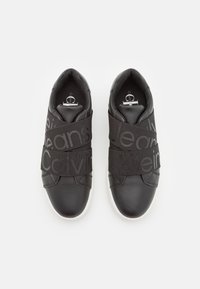 Zwarte sneakers met een gladde leren afwerking, voorzien van elastische banden met de tekst "Calvin Klein Jeans" en witte rubberen zolen.