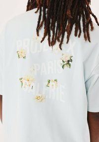 Camiseta de manga corta azul claro con detalles florales blancos y texto: "PROJECT X PARIS GALERIE" en blanco. Textura de tela suave y lisa.