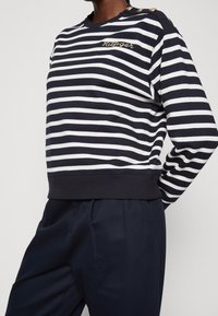 Sweatshirt rayé bleu marine et blanc avec un logo doré brodé et des boutons décoratifs sur l'épaule ; fabriqué en tissu doux et extensible.