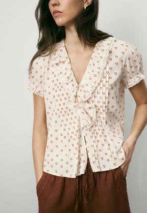 Woman with long dark hair wears cream blouse with red flower pattern, ruffled collar, and short sleeves, paired with brown pants, hands in pockets.