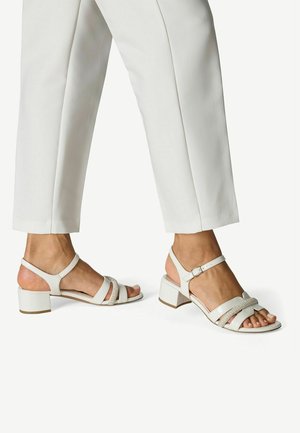 Sandales blanches à talons carrés avec plusieurs brides, texture en cuir lisse et accent métallique. Portées avec un pantalon blanc sur mesure.