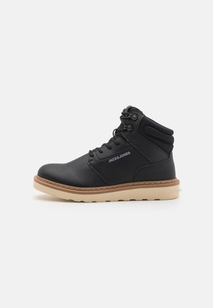 Botas altas negras, hechas de cuero sintético con un acabado texturizado, con suela de goma y suela exterior beige en contraste. Diseño con cordones.