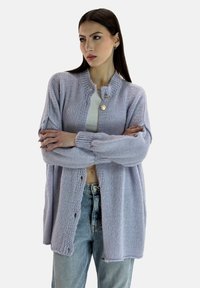 Cárdigan de punto lavanda claro con cuello redondo, detalles de botones y un corte holgado. Lleva puesto sobre una camiseta blanca y combinado con unos vaqueros azul claro.