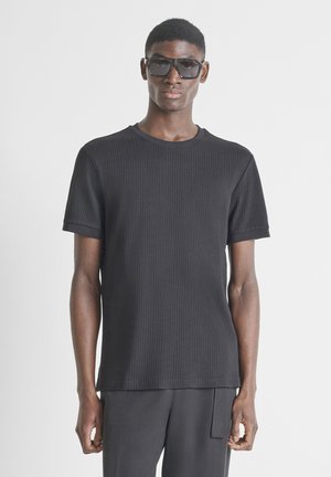 Giovane uomo in piedi, indossa una maglietta nera a maniche corte a coste, pantaloni neri e grandi occhiali da sole scuri su uno sfondo chiaro uniforme.