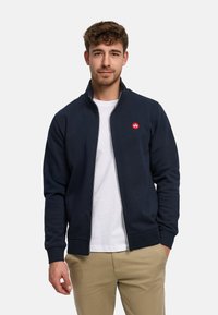 Mörkblå zip-up-tröja med hög krage, ribbade ärmslut och en liten röd logotyp på bröstet. Mjukt tyg med en slät yta.