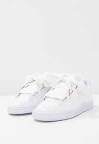 Bílé sportovní boty z umělého materiálu, s hladkým povrchem, silnými tkaničkami, zlatými detaily a strukturovanou šedou podrážkou.