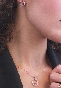 Set di gioielli in oro rosa composto da orecchini a bottone circolari e un collier con pendente e un accento di cristallo, esposto su uno sfondo scuro.