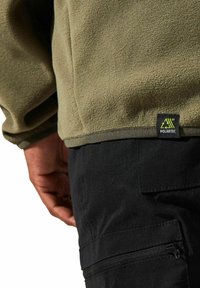 Olivenfarbener Fleece-Pullover mit strukturiertem Finish, ausgestattet mit einem Markenetikett am Saum "Polartec", kombiniert mit schwarzen Cargo-Hosen mit Reißverschlusstaschen.