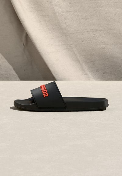 Μαύρο σανδάλι με φαρδιά λουράκια και κόκκινο λογότυπο "DSQUARED2" στη λουρίδα, τοποθετημένο σε ουδέτερη επιφάνεια με φόντο μπεζ ύφασμα.