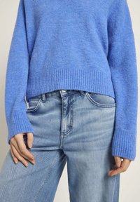 Blauer Strickpullover mit geripptem Saum und Bündchen, kombiniert mit hellen Jeans. Der Pullover hat eine lässige Passform und eine weiche Textur.