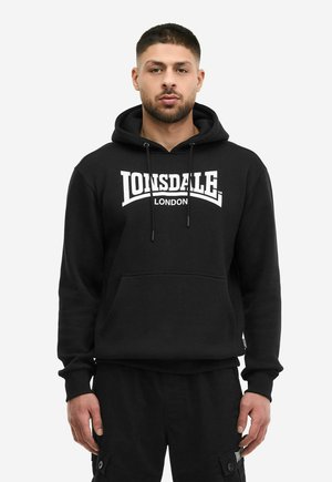 Mand iført sort Lonsdale London hoodie og sorte cargobukser, stående foran en ensfarvet hvid baggrund.