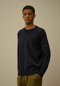 Maglione lavorato a maglia blu navy con scollo rotondo, polsini e orlo a coste, e un distintivo patch circolare sulla manica sinistra.