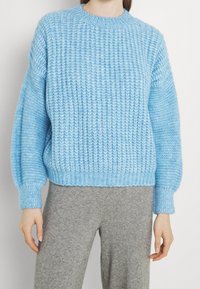 Suéter de punto azul claro con textura acanalada, cuello redondo y hombros caídos, contrarrestado con pantalones grises de textura.