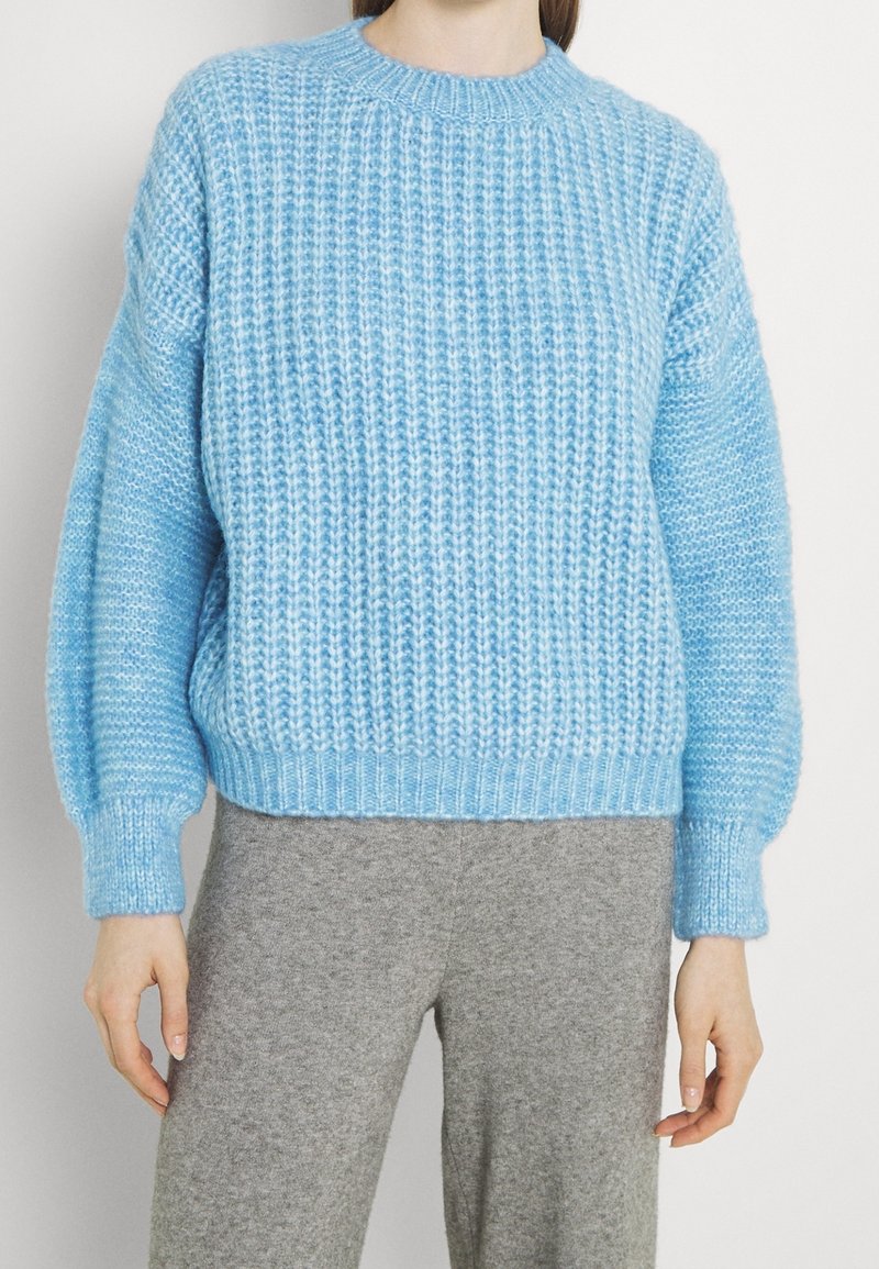 Suéter de punto azul claro con textura acanalada, cuello redondo y hombros caídos, contrarrestado con pantalones grises de textura.