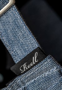 Correa de denim con un tejido azul texturizado, que presenta una etiqueta de tela negra con la palabra "Reell" en relieve en blanco. Hardware metálico plateado visible.