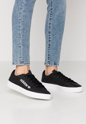 Nogi w jasnoniebieskich dżinsach, czarne buty sportowe Adidas z białymi podeszwami i logo z boku, stojące na jasnej podłodze.