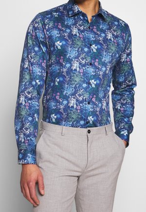 Mężczyzna w niebieskiej, kwiecistej, długorękawowej koszuli na guziki w połączeniu z jasnobeżowymi spodniami, ręce w kieszeniach, stoi na białym tle.