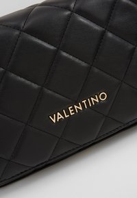 Musta tikattu nahkakotelo, jossa on kultainen "VALENTINO"-logo. Sujuva pinta, jossa on timantin muotoisia kuvioita ja hienoja ompeluyksityiskohtia.