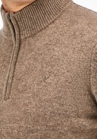 Brauner Strickpullover mit hohem geripptem Kragen, vertikalem Reißverschluss und subtiler Textur. Mit einem kleinen, geprägten Logo auf der Brust.
