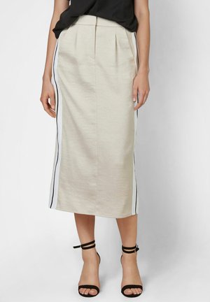 Frau trägt einen beigen Midirock mit weißen und schwarzen Seitenstreifen, kombiniert mit einem schwarzen Oberteil und schwarzen High Heels mit Knöchelriemen.