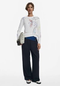 Witte trui met bloemenpatronen in roze en blauw, gecombineerd met brede zwarte broek. Accessoires omvatten een lichtgrijze tas en zwarte schoenen.