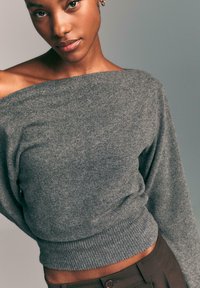 Szary sweter typu off-the-shoulder z ribbowanym wykończeniem i długimi rękawami, wykonany z miękkiej tkaniny, w zestawieniu z brązowymi spodniami o wysokim stanie.