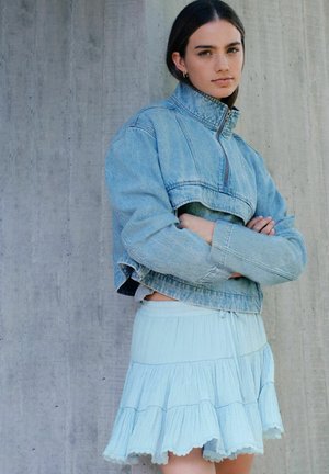 Kobieta ze skrzyżowanymi ramionami, ubrana w jasnoniebieską, krótką kurtkę dżinsową i pasującą do niej wielowarstwową spódnicę, stojąca na tle betonowej ściany.