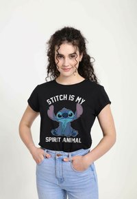 T-shirt noir avec un personnage de dessin animé aux tons bleus et gris, texte "Stitch est mon animal totem", manches retroussées et coupe décontractée.
