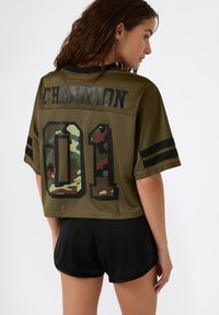 Oliwkowa krótka bluzka z jerseyu z nadrukiem "CHAMPION" i kamuflażowym wzorem "01" z tyłu. Krótkie rękawy z czarnymi paskami. W połączeniu z czarnymi szortami.
