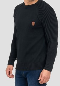 Pull en tricot noir avec manches et ourlet côtelés. Comprend un petit patch logo marron sur la poitrine. Design coupe régulière, matériau texturé.