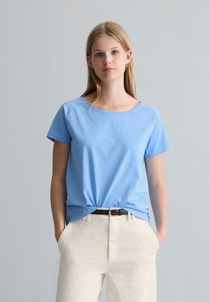 Giovane donna con lunghi capelli biondi che indossa una camicia azzurro chiaro a maniche corte infilata in pantaloni bianchi con le mani nelle tasche.