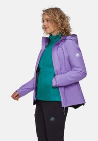 Chaqueta impermeable de color púrpura claro con capucha, sobre una capa intermedia aislante en color teal, combinada con pantalones negros que presentan un logo. Textura suave, diseño funcional.