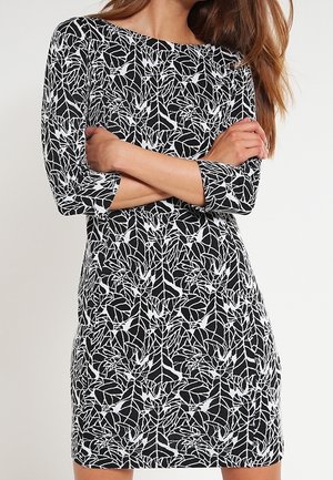 Femme aux longs cheveux portant une robe à motifs de feuilles noires et blanches avec des manches trois-quarts, debout les bras croisés.