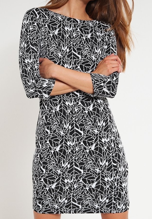 Femme aux longs cheveux portant une robe à motifs de feuilles noires et blanches avec des manches trois-quarts, debout les bras croisés.