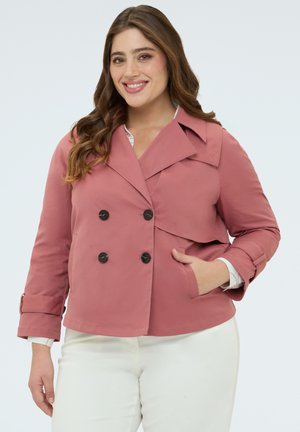 Donna sorridente con capelli lunghi castani che indossa una giacca doppiopetto color rosa polvere e pantaloni bianchi, mano in tasca, su sfondo chiaro.