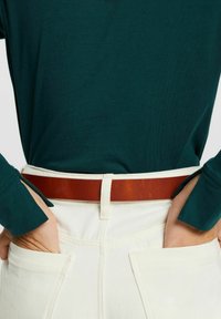 Dunkelgrünes langärmliges Oberteil, cremefarbene Hose und ein breiter brauner Ledergürtel. Das Shirt hat figurbetonte Ärmel und die Hose verfügt über Gesäßtaschen.