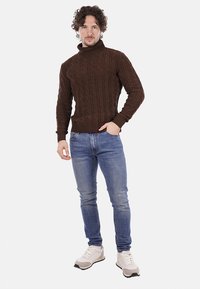 Maglione a collo alto in maglia marrone con motivo a trecce, abbinato a jeans azzurri chiari e sneakers bianche. La vestibilità è casual e aderente.