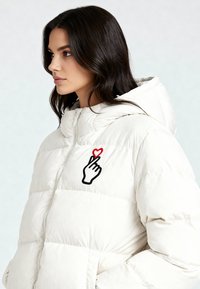 Giacca a vento bianca con chiusura a zip, con grafica di una mano nera e un cuore rosso sul lato sinistro. Finitura trapuntata testurizzata.