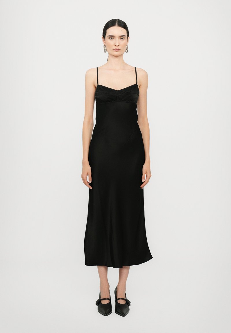 Vestido de satén negro con tirantes finos, cuerpo ajustado y falda fluida. Combinado con zapatos negros de punta con lazos.