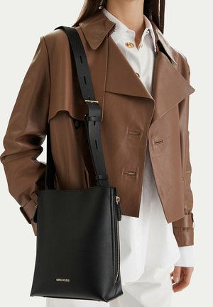 Kobieta w brązowej skórzanej kurtce i białej koszuli, niosąca czarną torebkę na ramię marki Gino Rossi z szerokim regulowanym paskiem.
