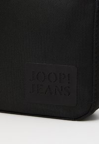 Detail čiernej textúrovanej látky s čiernou nášivkou s embosovaným logom "JOOP! JEANS" v rohu tašky.